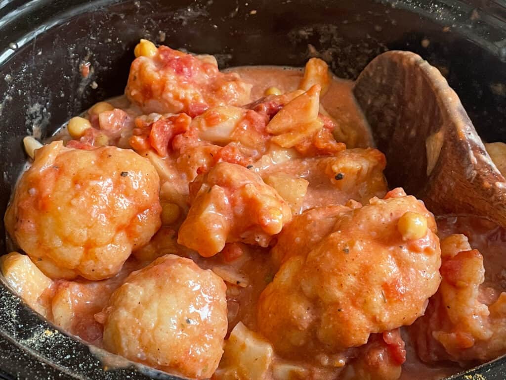 Cauliflower mixed with all ingredients in slow cooker, with wooden spoon.