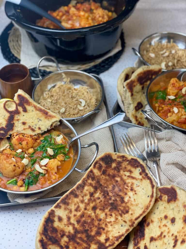 Naan breads in bowl, two silver bowls of curry, two smaller bowls or rice, slow cooker to the side, cream and white background.