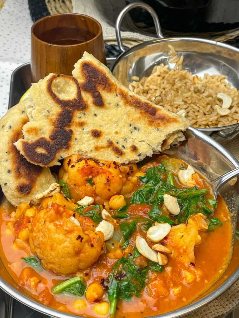 Cauliflower tikka masala in small silver curry bowl, with naan bread at side, small silver curry dish with brown rice, small brown cup to side.