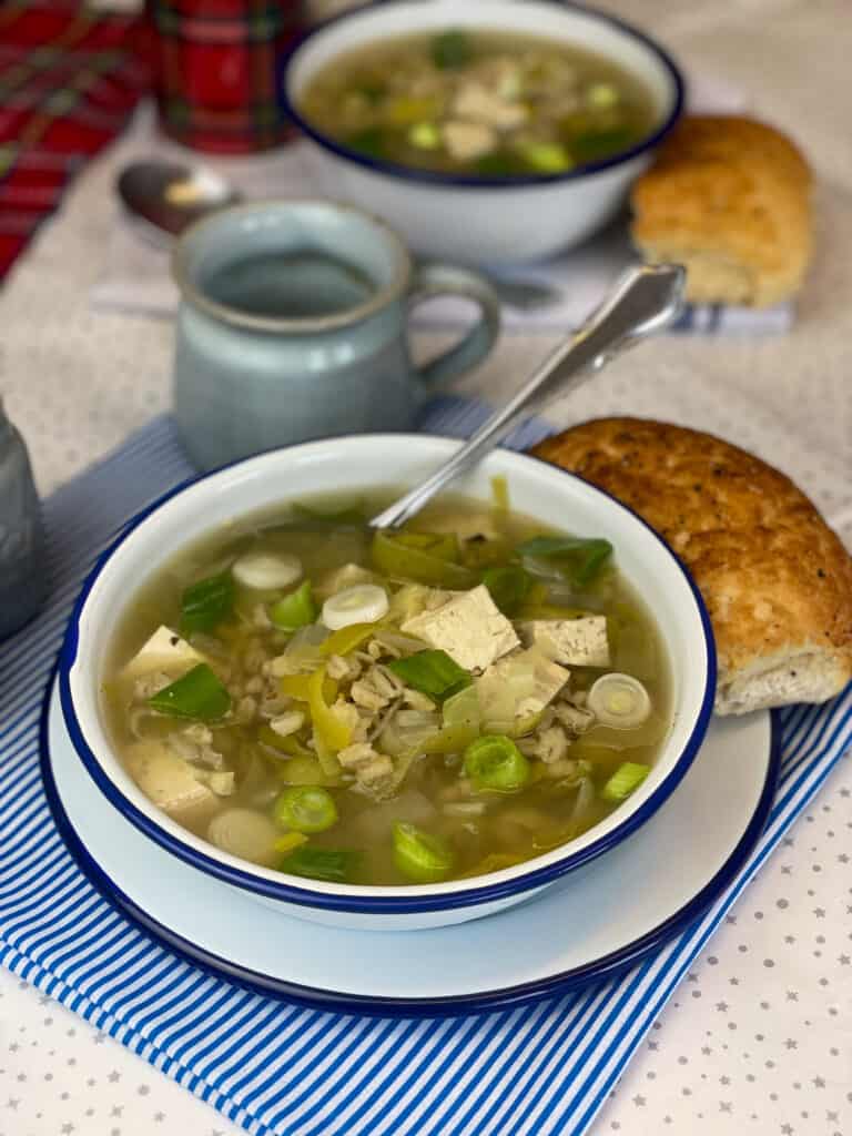 white bowl with blue rim full of cock-a-leekie soup with bread roll to side and small blue cup to side and second bowl of soup and roll in distance.