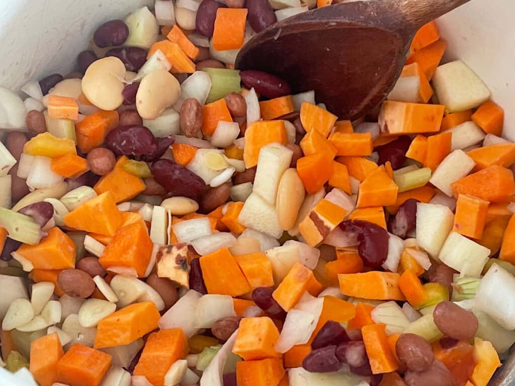 veggies and beans added to soup pan with wooden spoon.