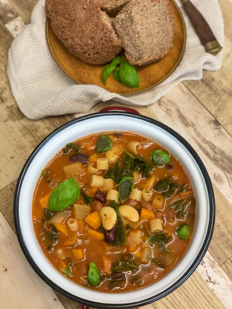 Bowl of Minestrone soup with brown wood plate with crusty bread, light brown wood background.