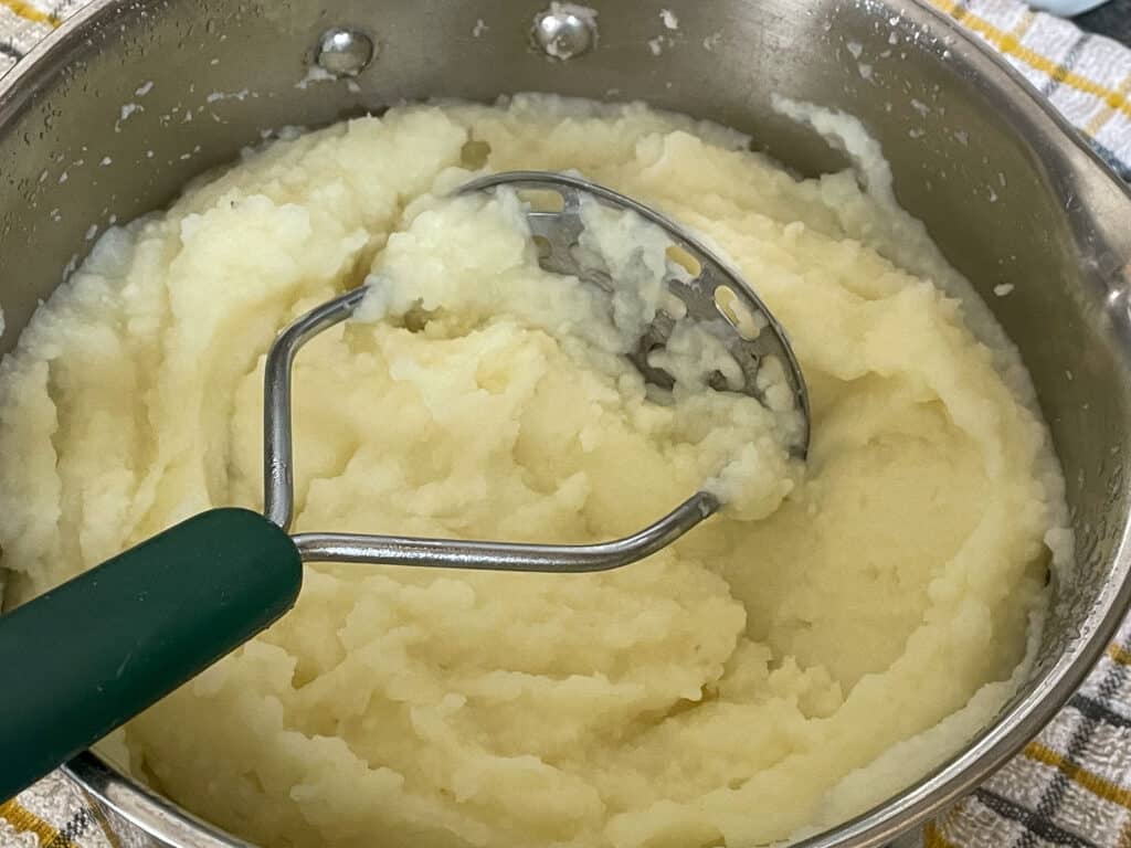 Mashed potatoes in saucepan with potato masher.