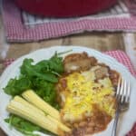 Vegan cowboy pie served with corn cobs and wild rocket in white bowl, silver fork, and red casserole dish in distance, red check tea towels.