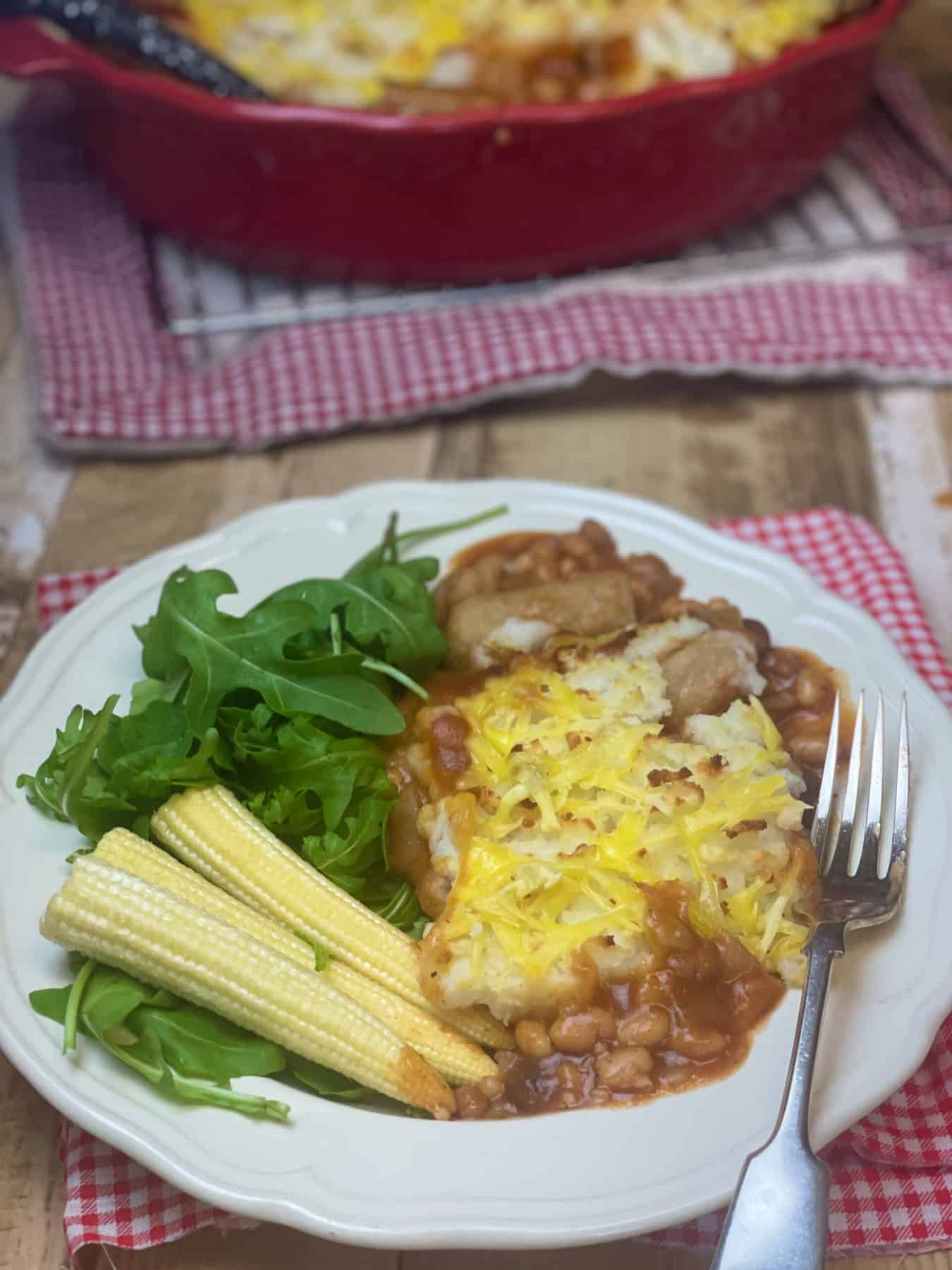Vegan cowboy pie served with corn cobs and wild rocket in white bowl, silver fork, and red casserole dish in distance, red check tea towels.
