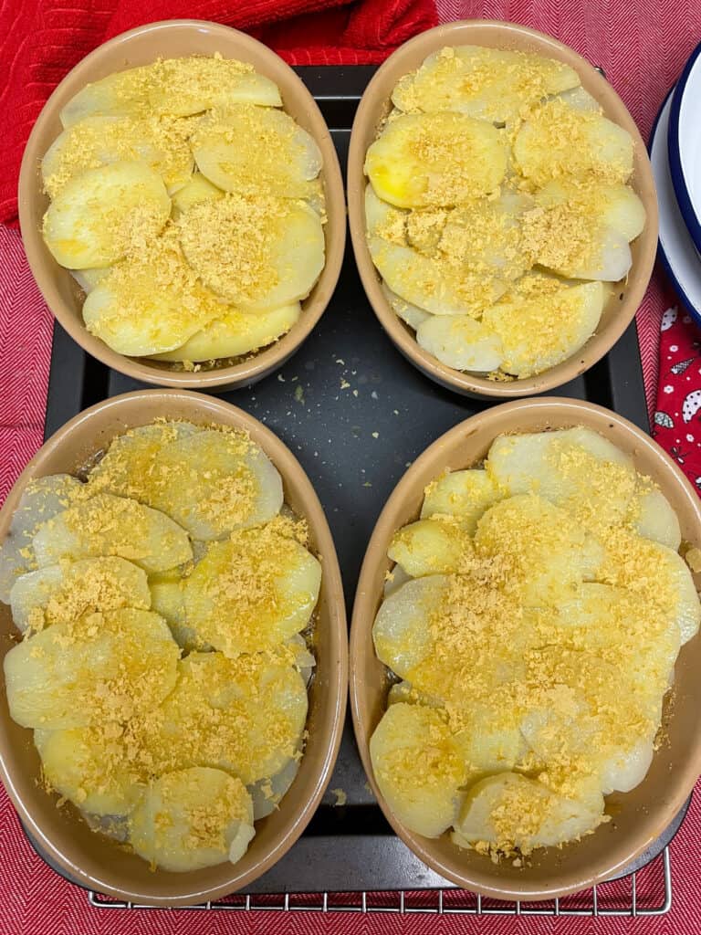 Potato slices added to hotpot mix, nutritional yeast flakes sprinkled on top, sitting on baking tray with red background.