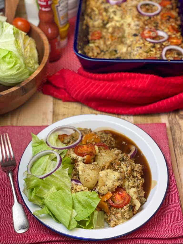 veggie hamburger casserole dished up on white plate with side salad and onion gravy, red napkin, salad bowl to side and casserole dish above.