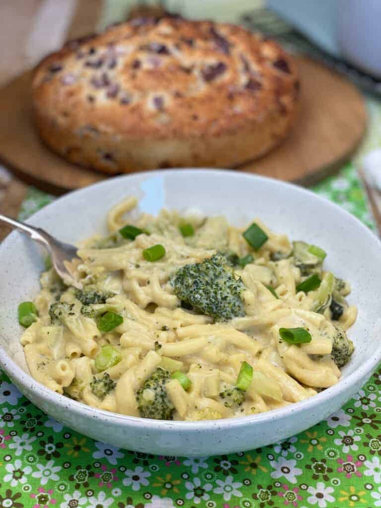 Served bowl of cheesy pasta with spring onion garnish and crusty bread in background.