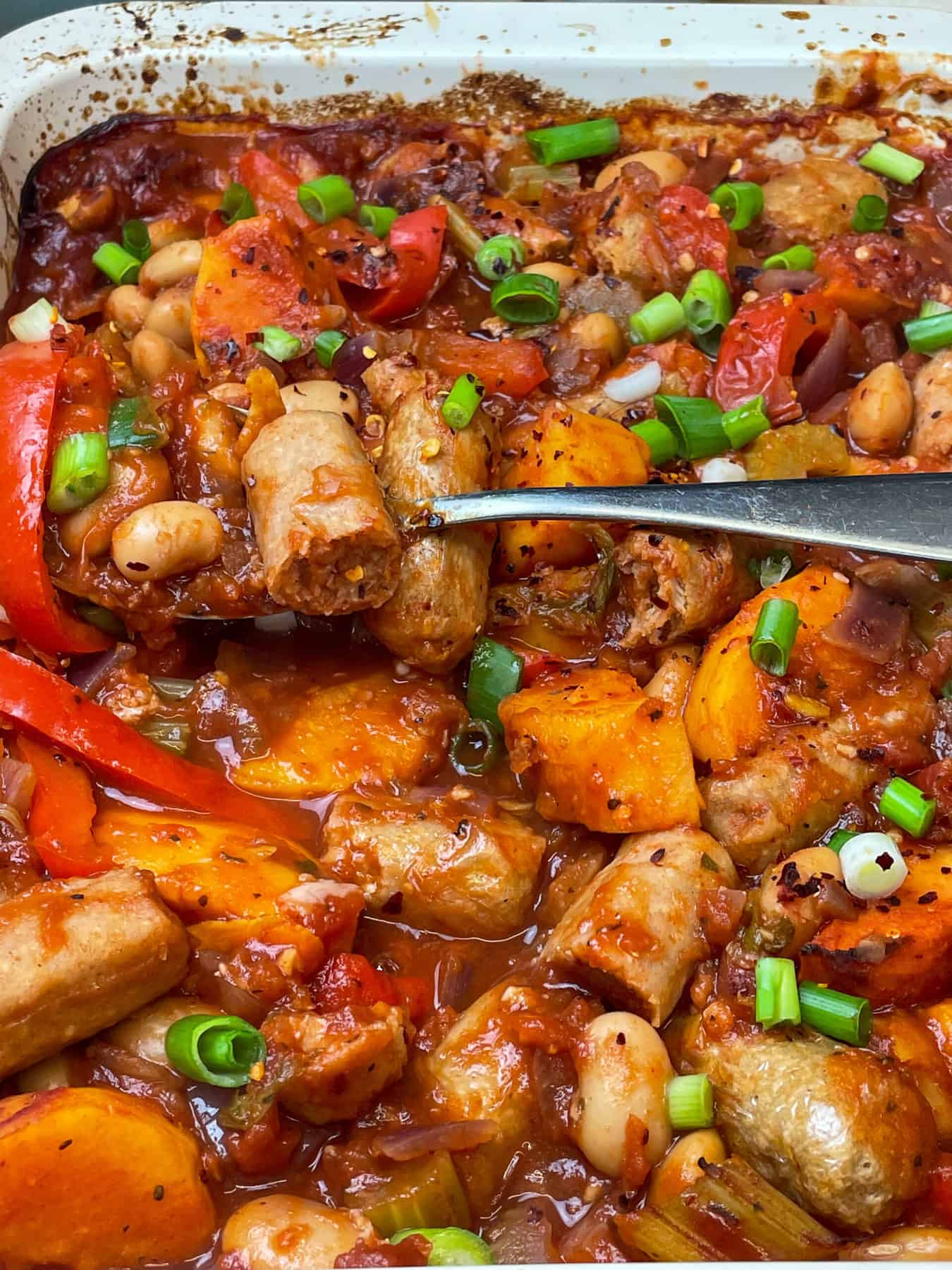 Vegan sausage casserole garnished with spring onions and a silver spoon scooping out a serving.