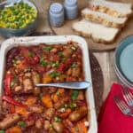 Tray of sausage casserole with sliver serving spoon, bowl of peas and corn to side, slices of bread on breadboard, blue salt and pepper shakers, red napkin with forks to side.