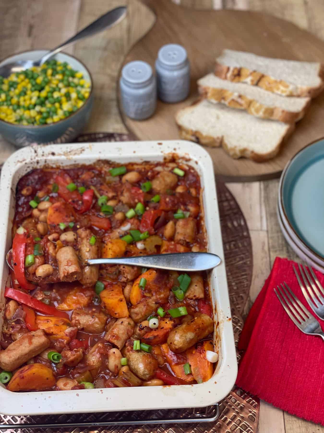 Tray of sausage casserole with sliver serving spoon, bowl of peas and corn to side, slices of bread on breadboard, blue salt and pepper shakers, red napkin with forks to side.