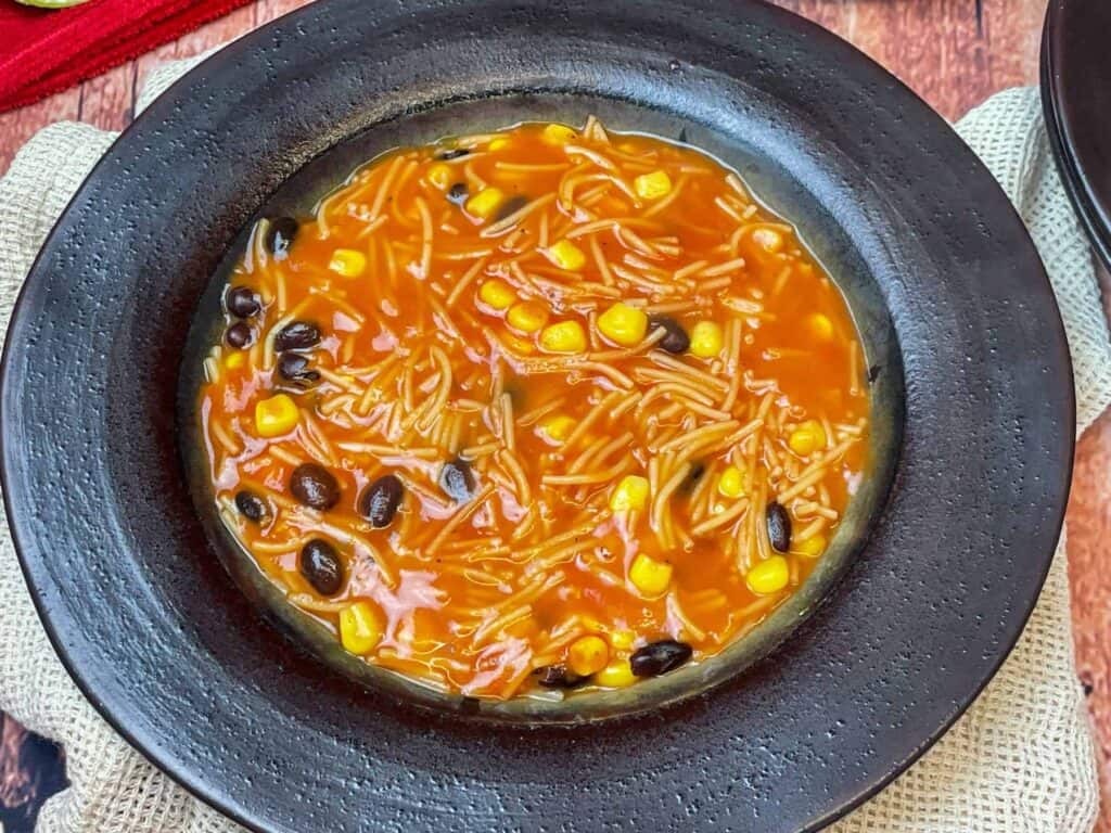 Mexican noodle soup served in dark brown bowl with red and cream background.