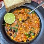 A dark brown clay bowl with Mexican noodle soup, lime wedge and silver spoon in bowl, and wedge of crusty bread to side.