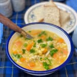 Vegan traditional Scotch broth in white blue rimmed enamel bowl with wooden handled spoon, small flower patterned plate with bread to side, blue salt and pepper shakers, blue tartan background.