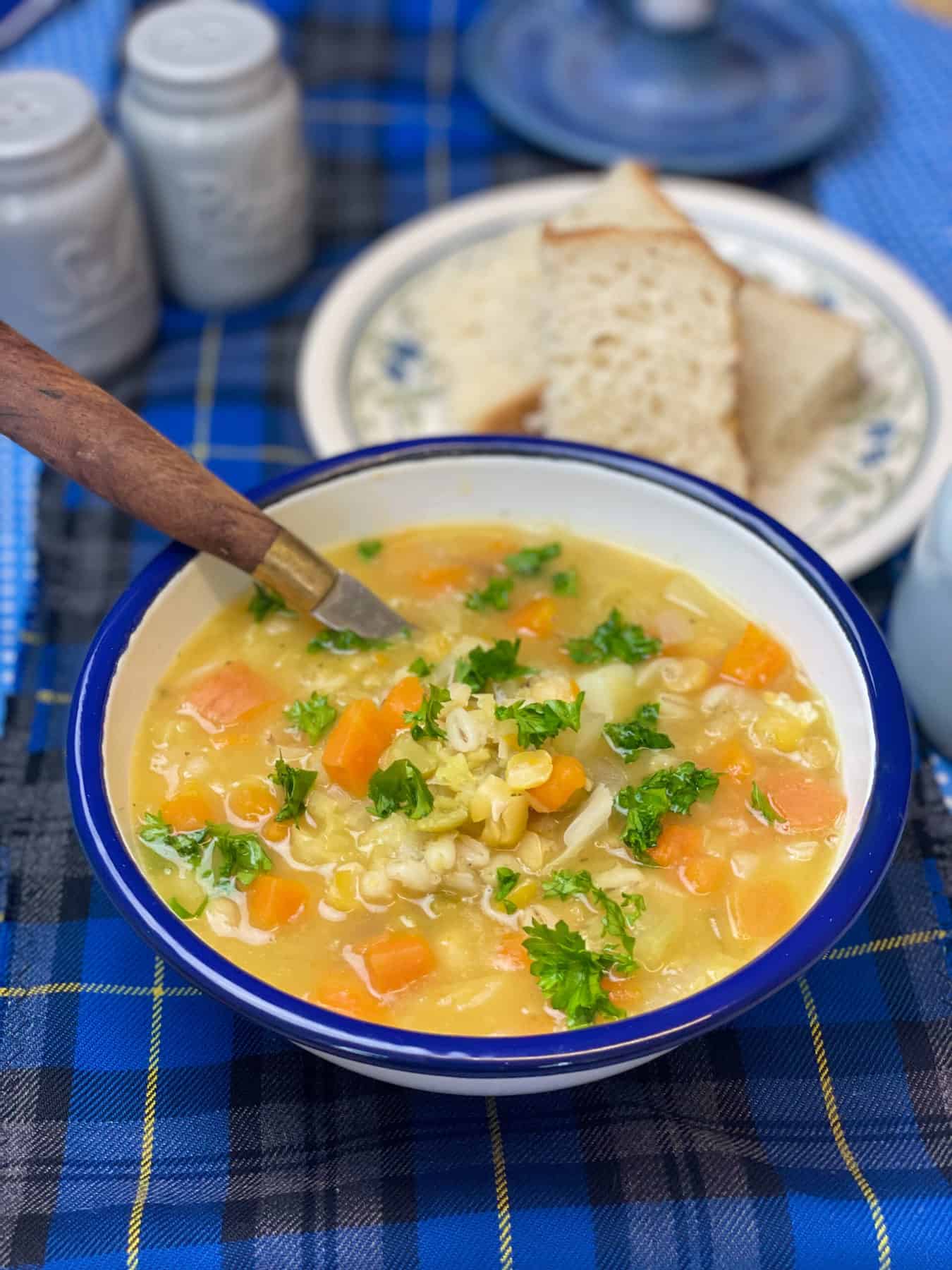 Vegan traditional Scotch broth in white blue rimmed enamel bowl with wooden handled spoon, small flower patterned plate with bread to side, blue salt and pepper shakers, blue tartan background.