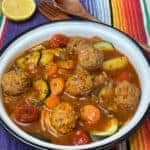 Bowl of Mexican meatball soup with lemon wedge, wooden fork and knife, and colourful stripy background.