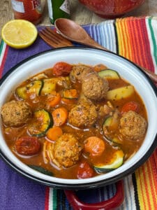 Bowl of Mexican meatball soup with lemon wedge, wooden fork and knife, and colourful stripy background.