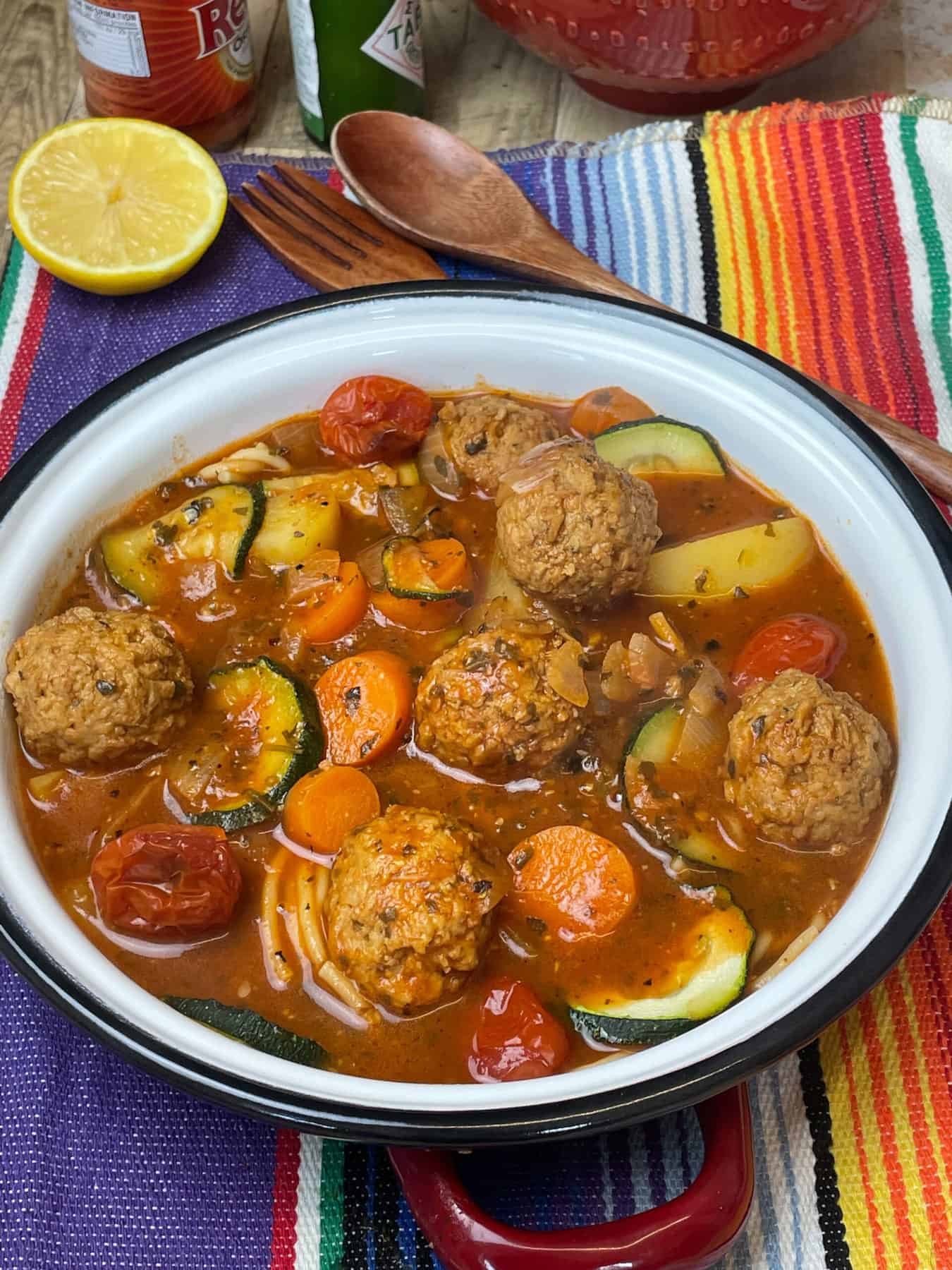 A white bowl with black rim filled with Mexican meatball soup, stripy background with lemon.