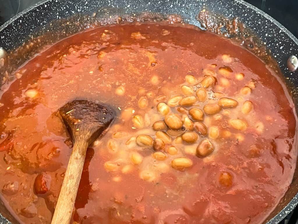 Pinto beans added to pasta sauce with wooden spoon.