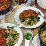 Two bowls of vegan sausage pasta with skillet to side, and wooden background, small dish of chopped herbs.