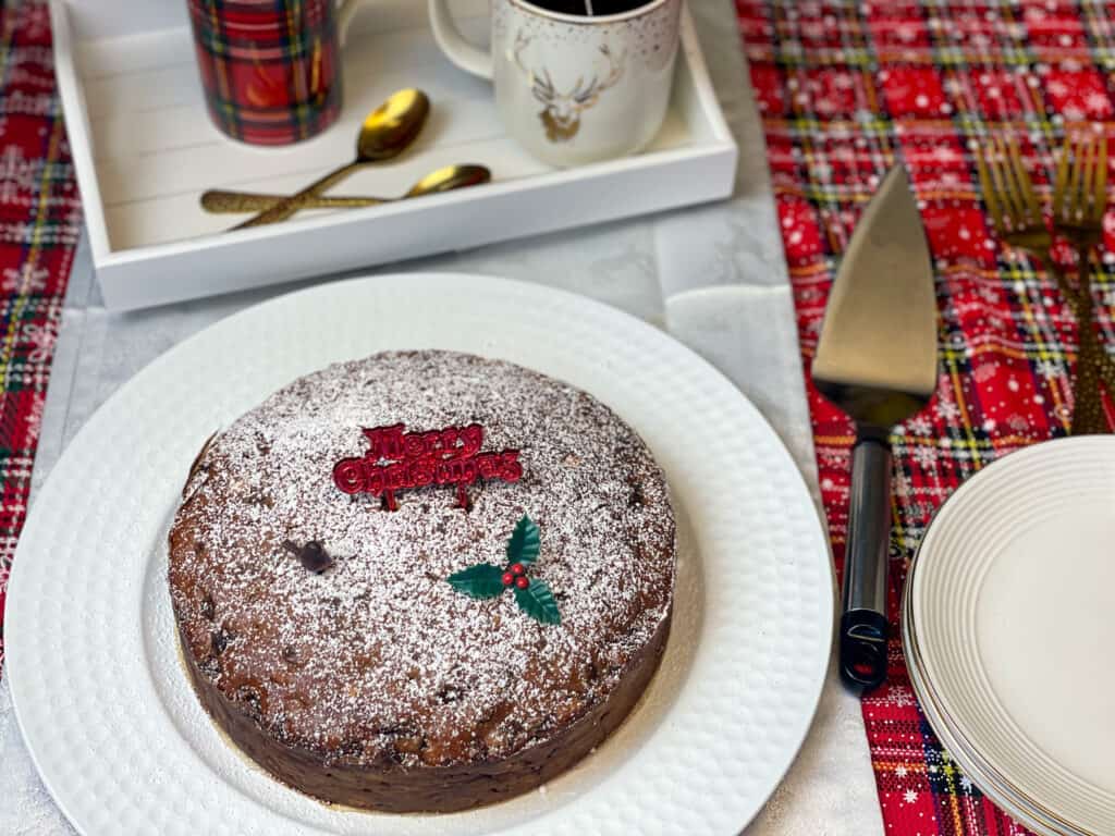 3 Ingredient vegan Christmas cake served with dusting of icing sugar and cake decorations, cake slice to side, and tea tray to side.