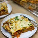 Plate with lasagna slice, fork and knife, second plate to side and baking dish to side, tan background.