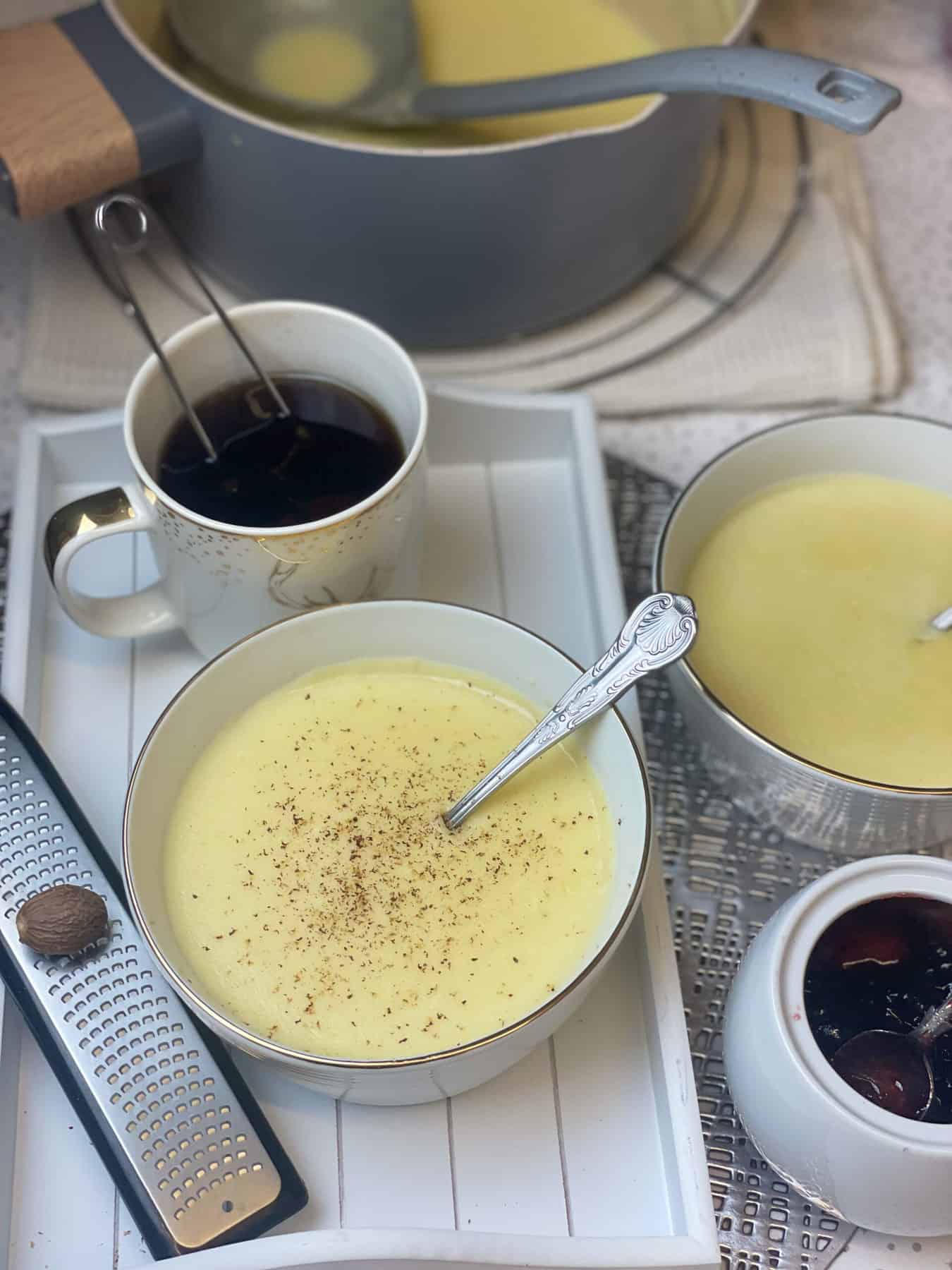 Featured image, tray with semolina pudding and mug of tea, nutmeg and zester to side, saucepan with semolina to back, jam pot to side and second bowl of pudding to side.