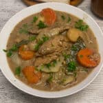 A white bowl of vegan creamed beef and barley stew with parsley, plate with bread to side, featured image.