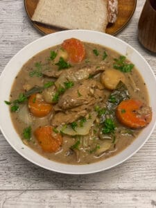 A white bowl of vegan creamed beef and barley stew with parsley, plate with bread to side, featured image.