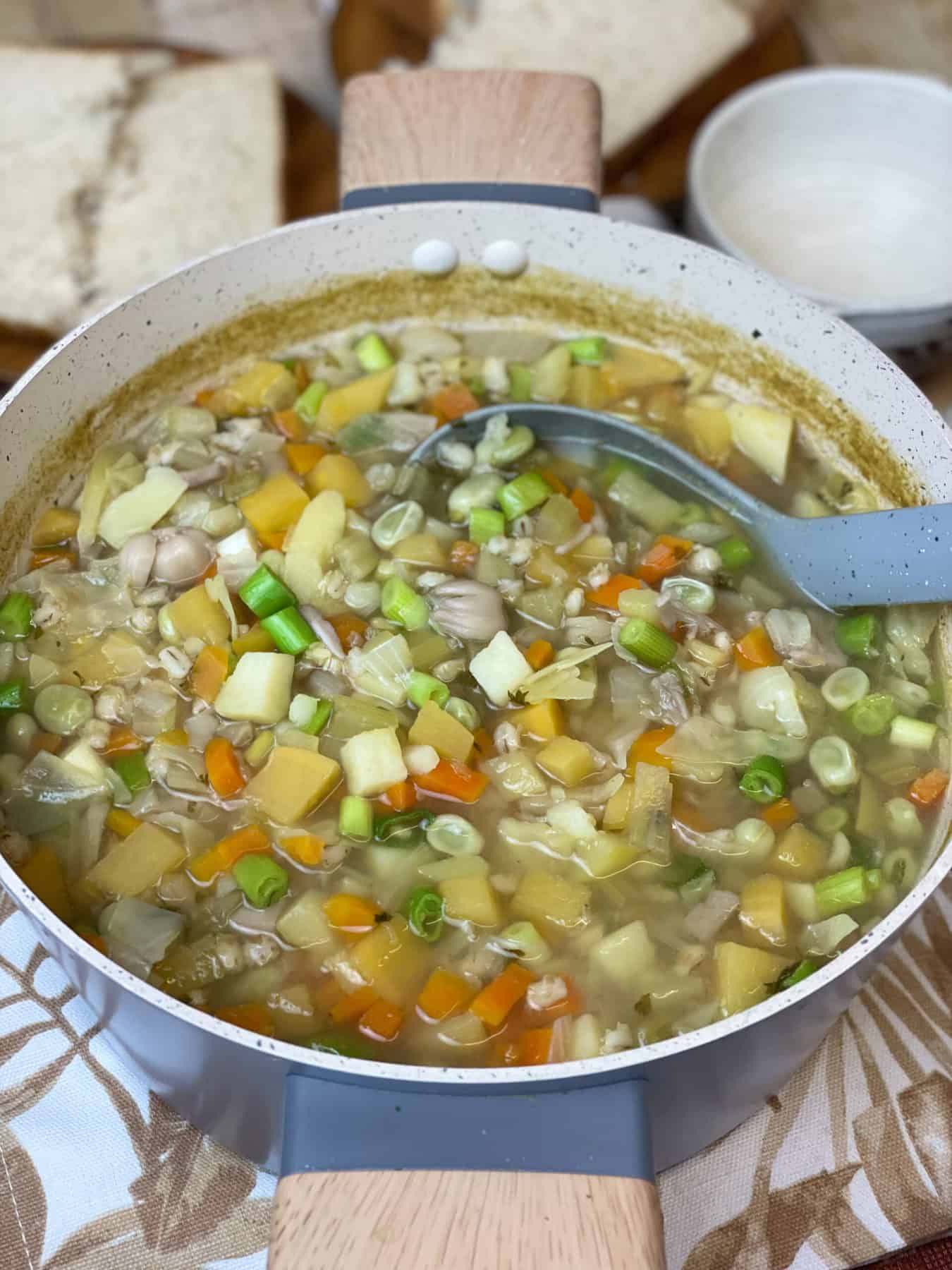 Close up of soup pot with wooden handles full of hotchpotch soup, with grey ladle and small soup bowl to side.