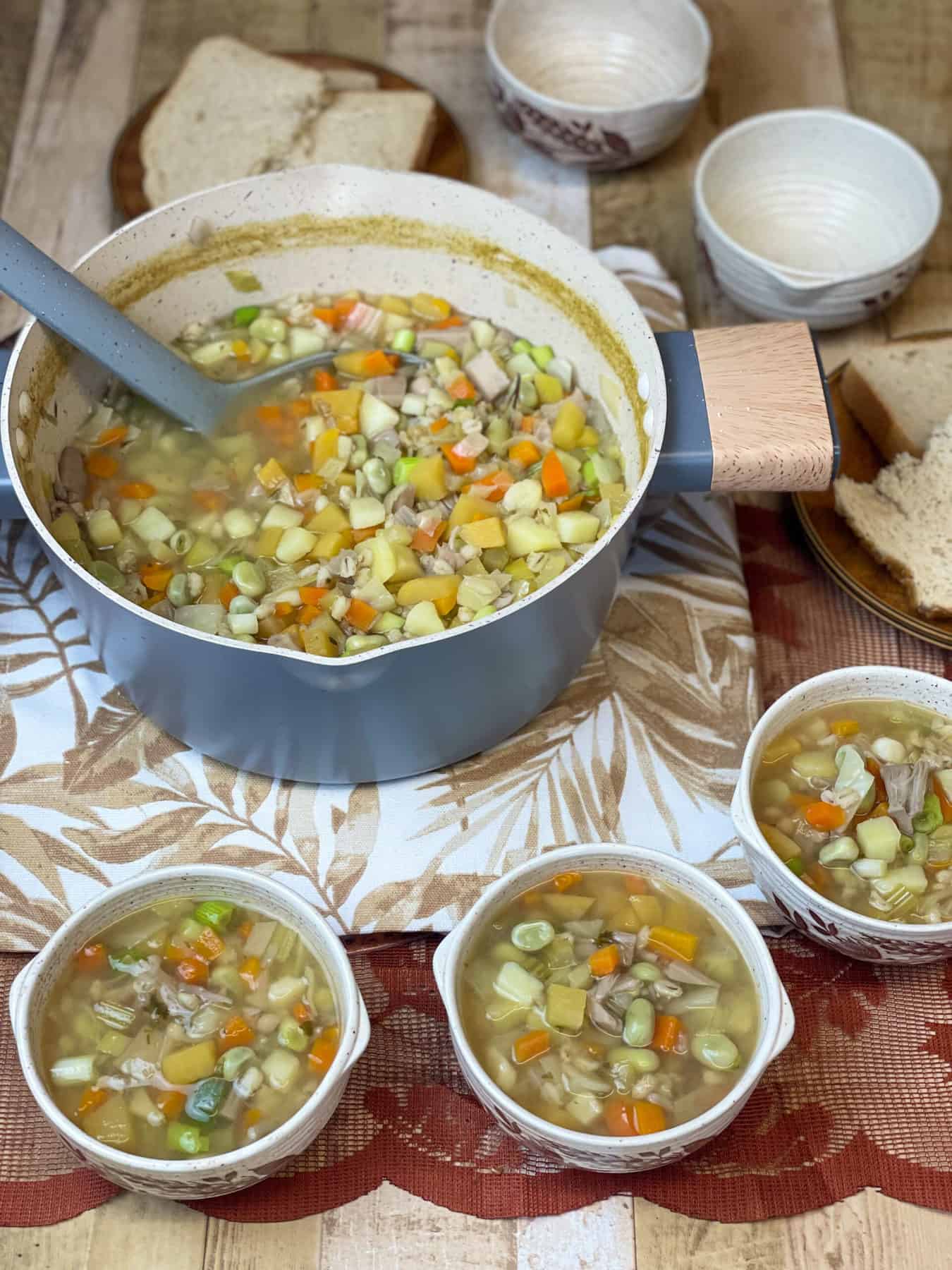 A pot of Scottish Hotchpotch with three bowls full to side, and two empty bowls, brown leaf tea towel background featured image.