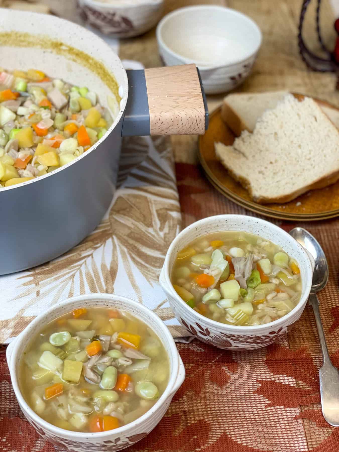 Hotchpotch pot to side with two small bowls filled with soup, bread to background, and a brown leaf table cloth.