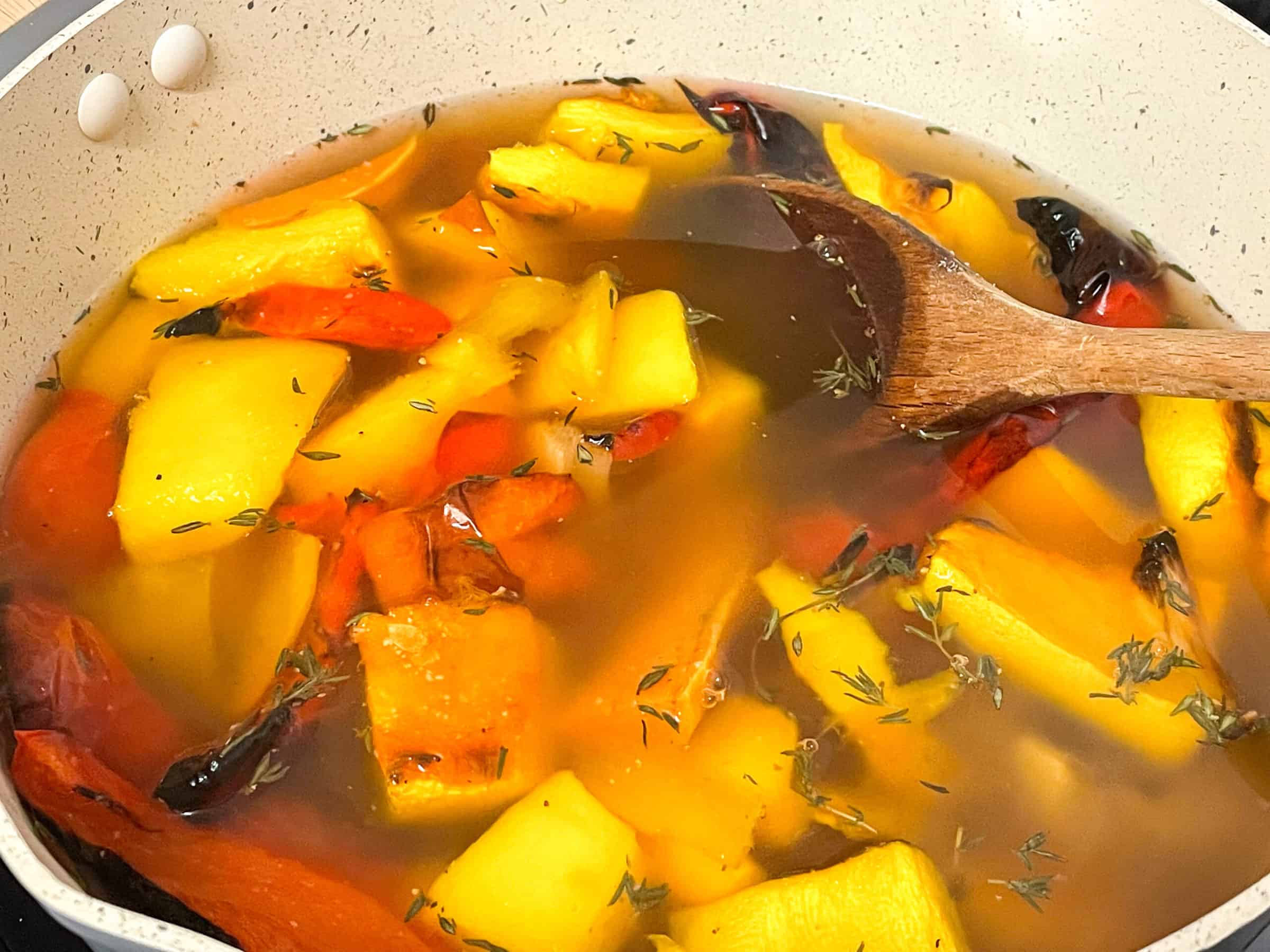Roasted veggies added to soup pan along with veggie stock, wooden spoon to side.