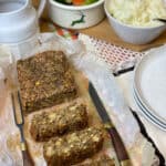 vegan traditional meatloaf with chickpeas, green lentils and oatmeal on chopping board, with knife, and bowl of mashed potatoes, small pot of Brussel sprouts and carrots, and gravy dish to side, featured image.