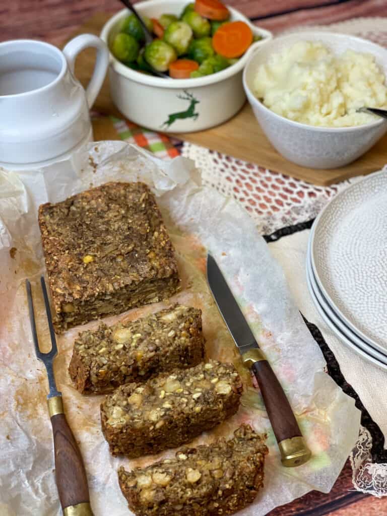 vegan traditional meatloaf with chickpeas, green lentils and oatmeal on chopping board, with knife, and bowl of mashed potatoes, small pot of Brussel sprouts and carrots, and gravy dish to side, featured image.