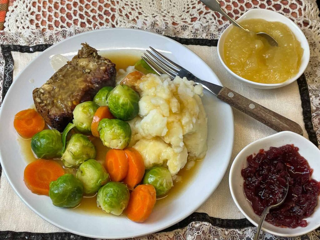 vegan sage and onion meatloaf served on white plate with Brussel sprouts, carrots, mashed potatoes, vegan gravy and small dishes of cranberry and apple sauce to side, wooden handle fork and lacy placemat.