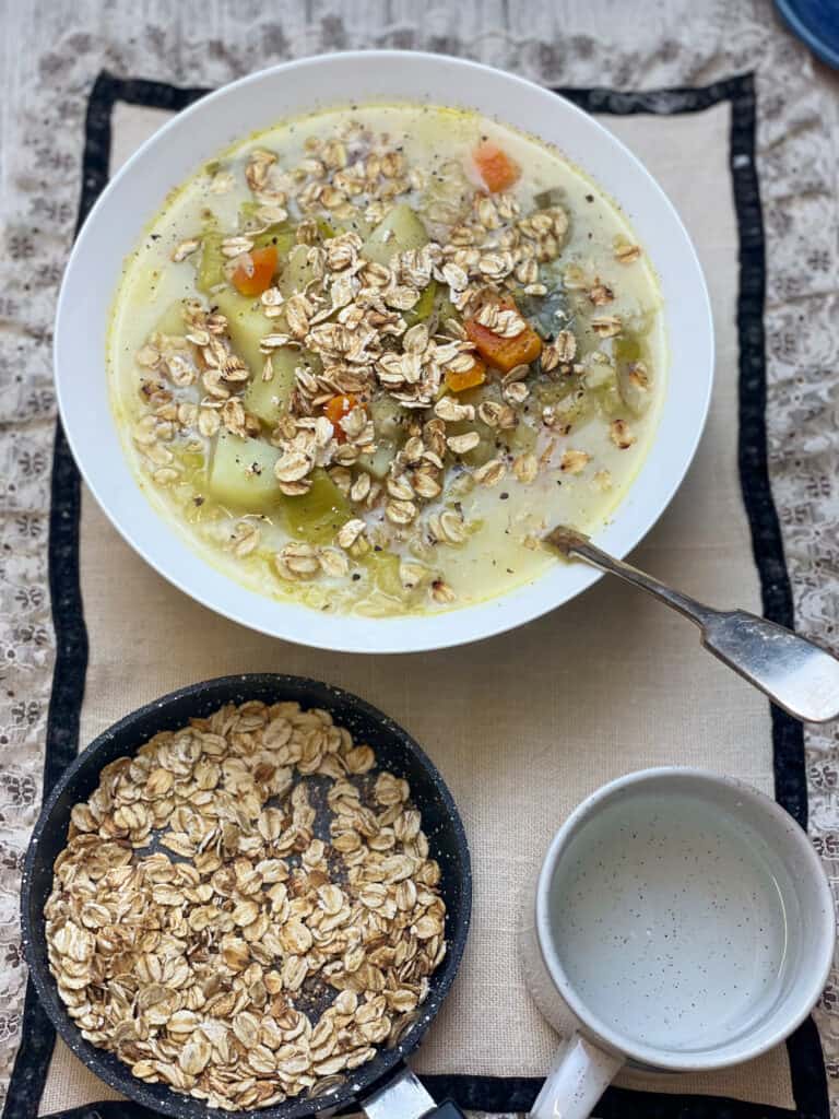 Scottish vegan traditional leek and potato soup in white bowl with small pan of toasted oats to side, water cup to side, and frilly placemat, featured image.