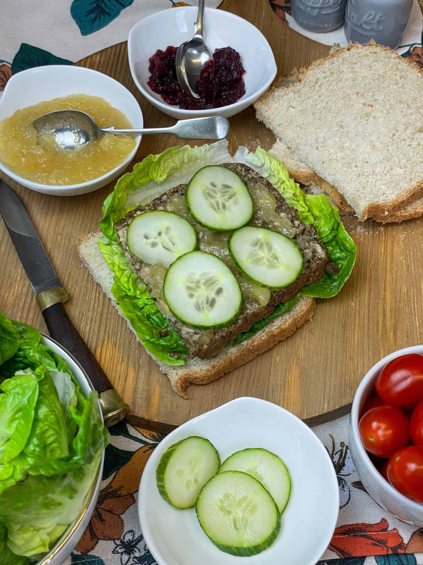 slices of bread with sliced vegan meatloaf on top, along with sliced cucumber and lettuce, apple and cranberry sauce dishes to side, salad dishes to side, all on wooden chopping board.