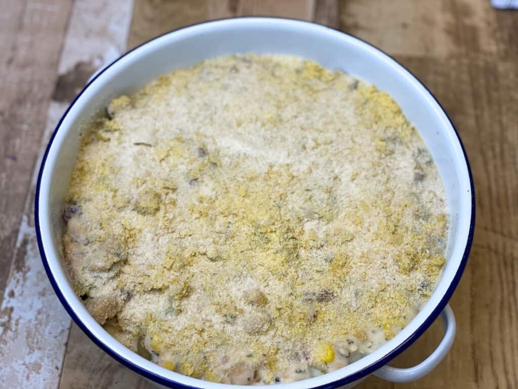 chickpea tuna and creamy sauce added on top of the potatoes and spinach, with nutritional yeast flakes and breadcrumb topping, in circular casserole dish ready to be baked.