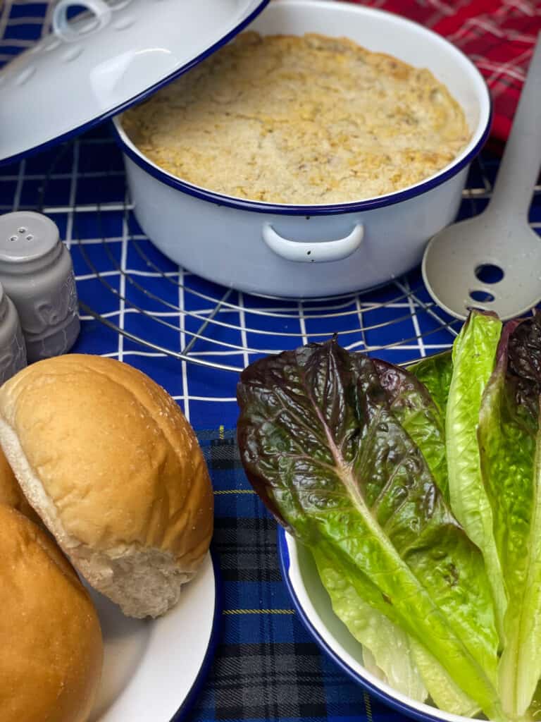 vegan chickpea tuna and potato mornay in casserole dish with lid to side, plate of bread rolls and plate of lettuce leaves, serving spoon to side, blue and white check tea towel background.