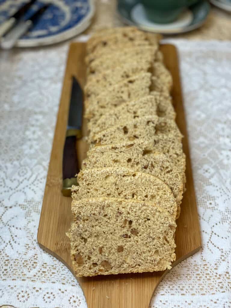 peanut butter bread sliced and sitting on long wooden chopping board with knife to side, tea cup and saucer to top and blue and white serving plate with cutlery to side, white Dollie placemat background.