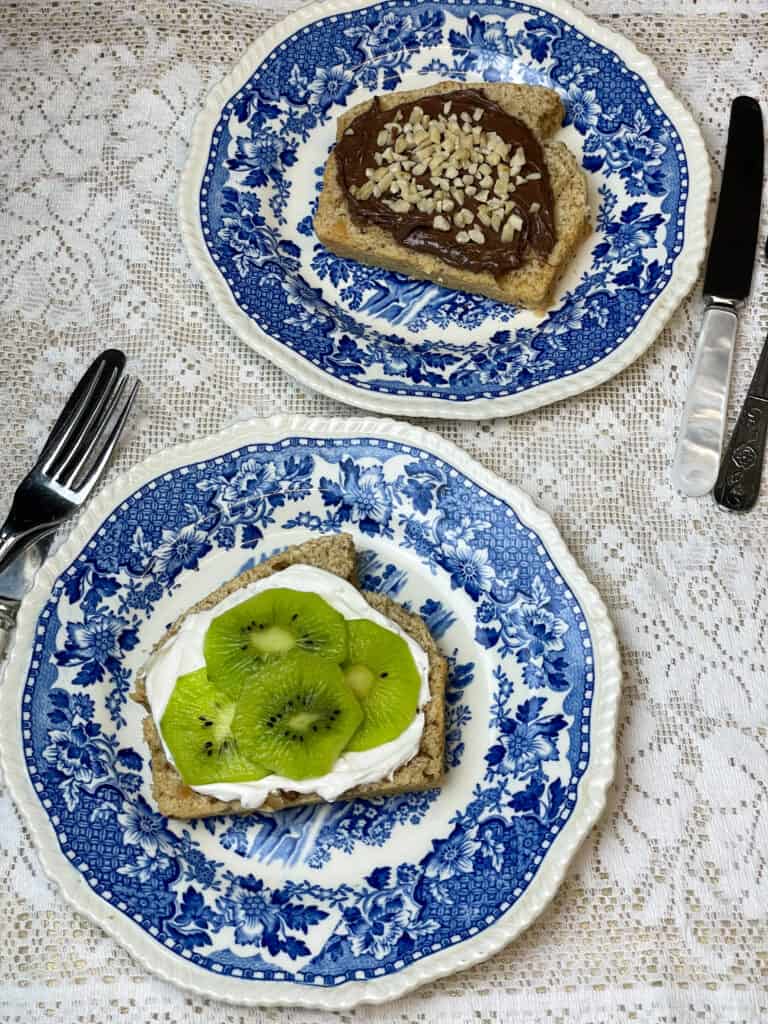 two plates with bread slice, one slice has cream cheese and kiwi topping, the other slice has chocolate spread and mixed nut topping, dessert knifes to side, white lacy background.