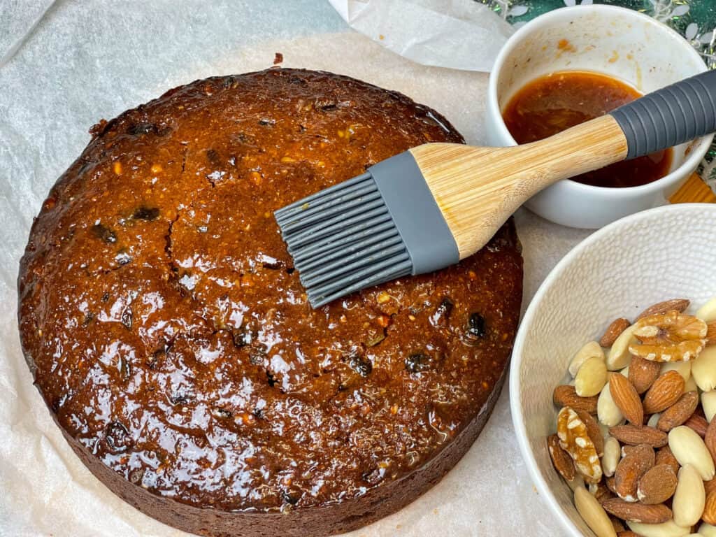 undecorated vegan Christmas cake with pastry brush on top glazing the surface with a fruit glaze, small glaze bowl to side, larger bowl with whole nuts in to the side, greaseproof paper background.