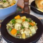 A brown bowl filled with vegetable stew and dumplings, brown salt mill to background with blue stew pot, and plate of bread.