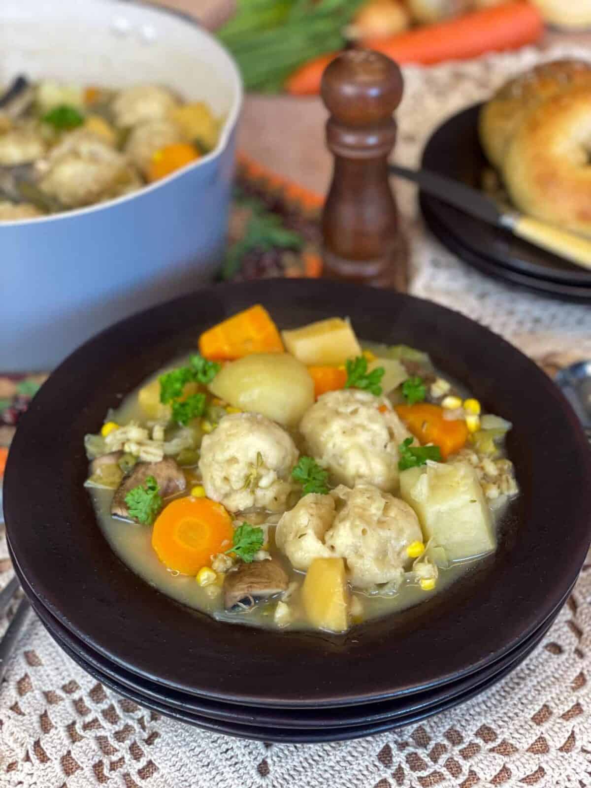 A brown bowl filled with vegetable stew and dumplings, brown salt mill to background with blue stew pot, and plate of bread.