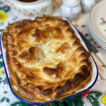 A vegan chicken pie on a green trivet with flower patterned gravy jug in background with plates to side, bright colourful flower tablecloth.