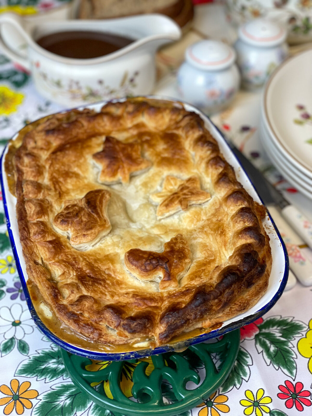 A vegan chicken pie on a green trivet with flower patterned gravy jug in background with plates to side, bright colourful flower tablecloth.