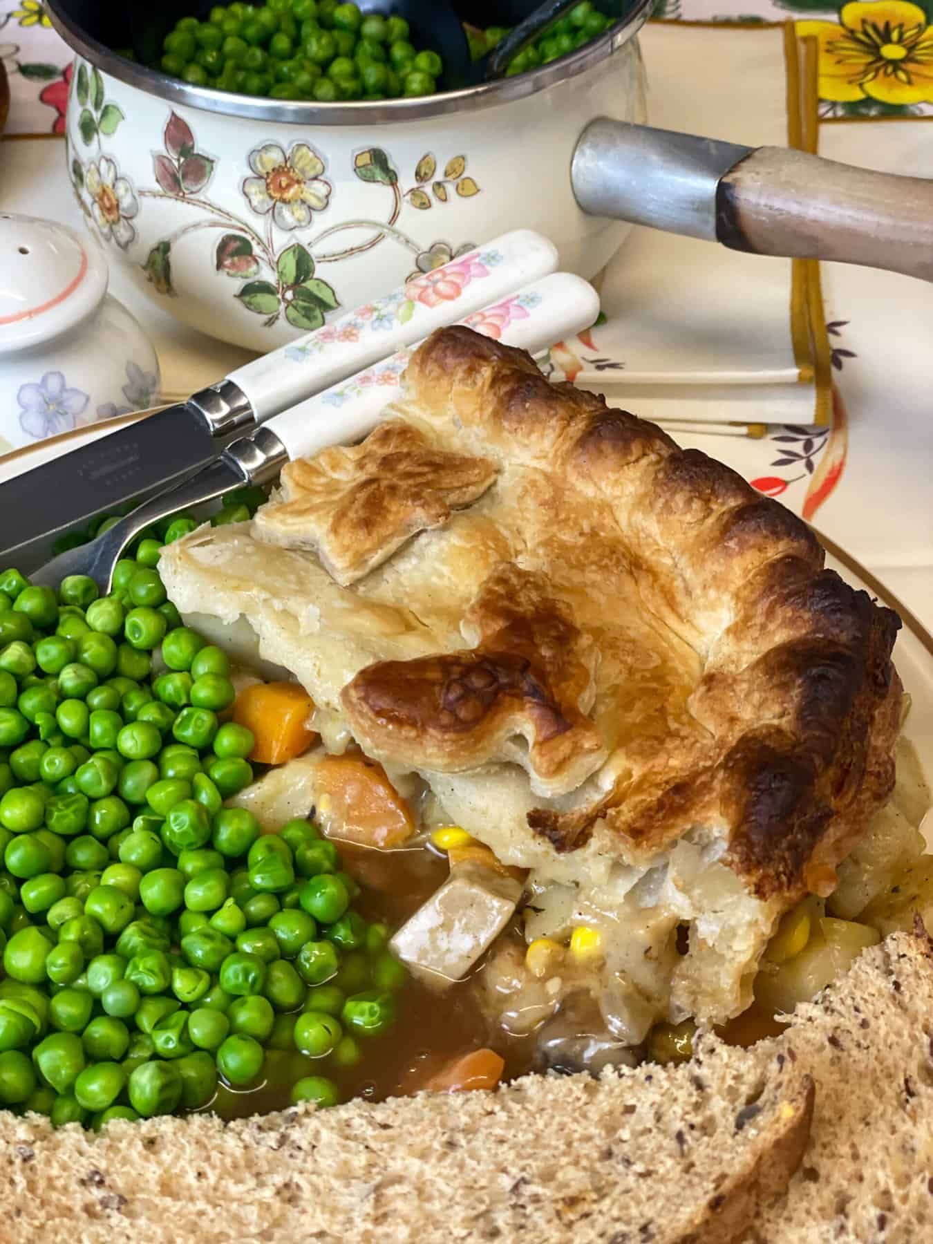 Slice of vegan pie with peas, bread, gravy on dinner plate, flower patterned pot of peas in background, with flower handled fork and knife.