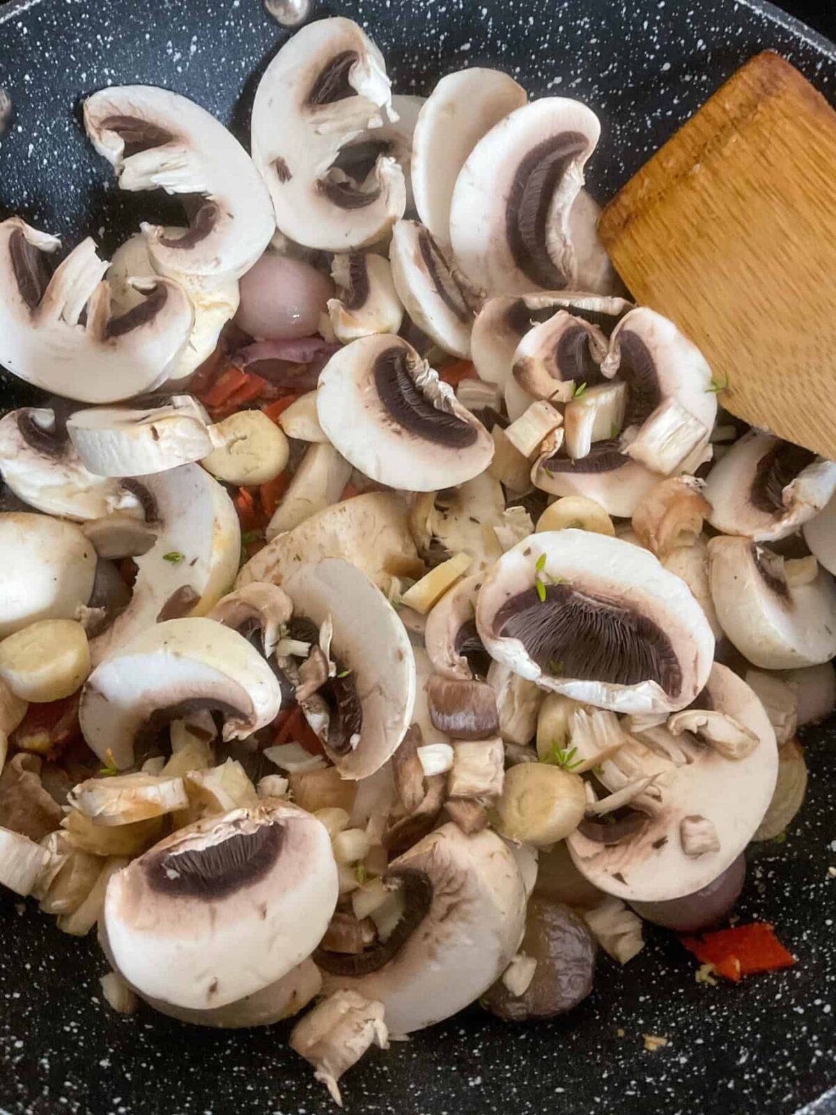 Sliced mushrooms added to pan with shallots and vegan bacon, wooden spatula to side.