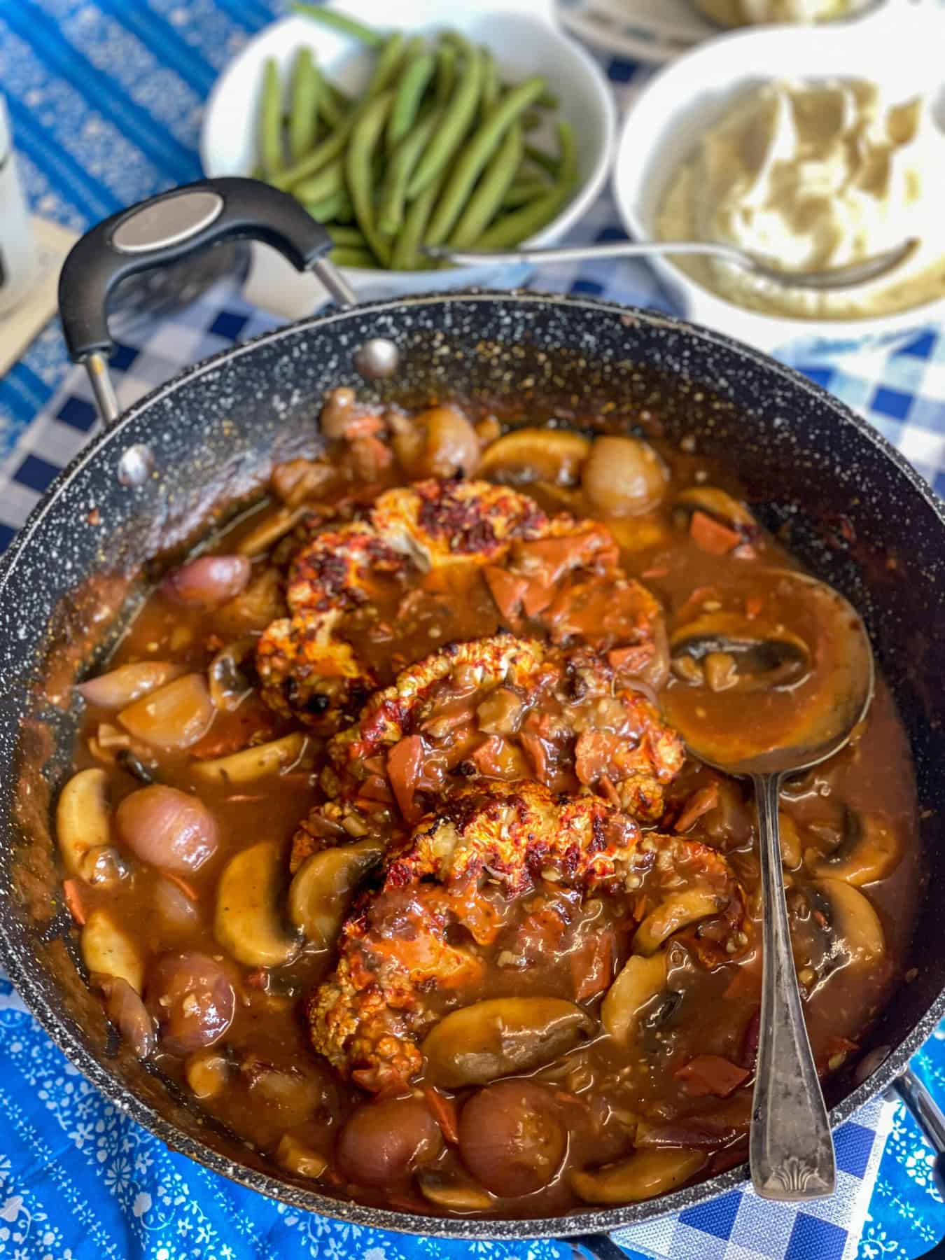 Skillet filled with cauliflower steaks and coq au vin sauce, side dishes of green beans and mashed potatoes in background, blue and white stripe table cloth.